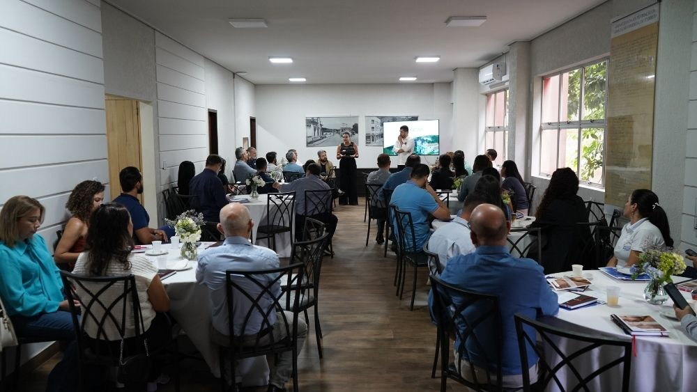 Palestrante ao centro da imagem, público sentado em mesas redondas nas laterais