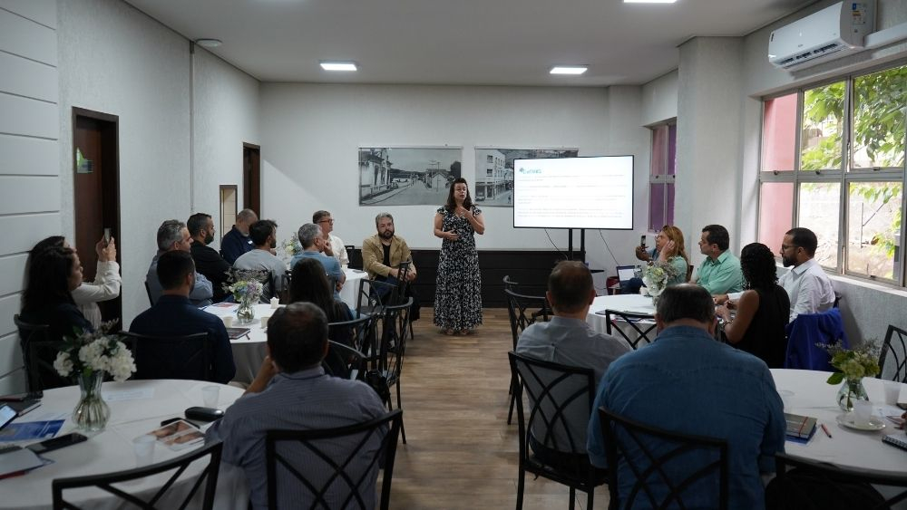 Palestrante ao centro e várias pessoas sentadas em mesas redondas nas laterais 